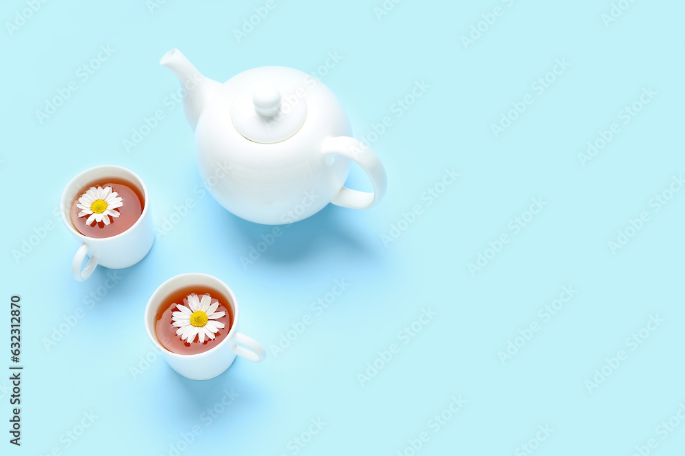 Teapot with cups of natural chamomile tea and flowers on blue background