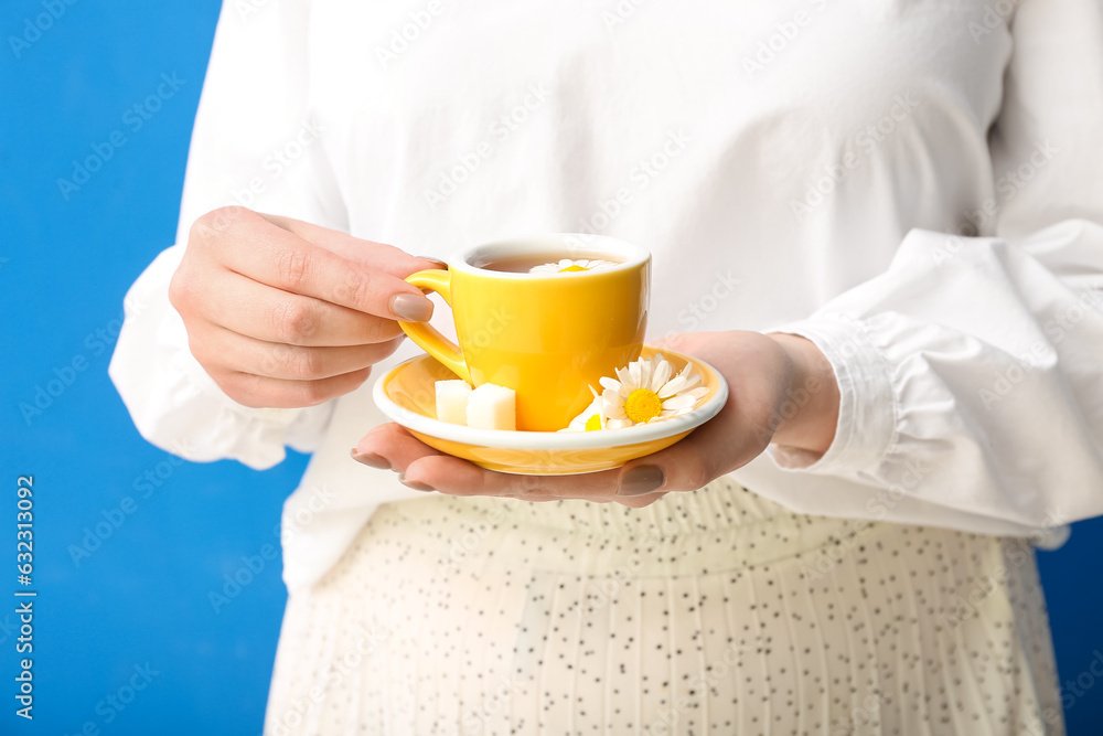 Beautiful young woman with cup of chamomile tea and flowers near blue wall