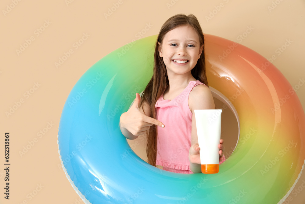 Little girl with inflatable ring pointing at bottle of sunscreen cream on beige background