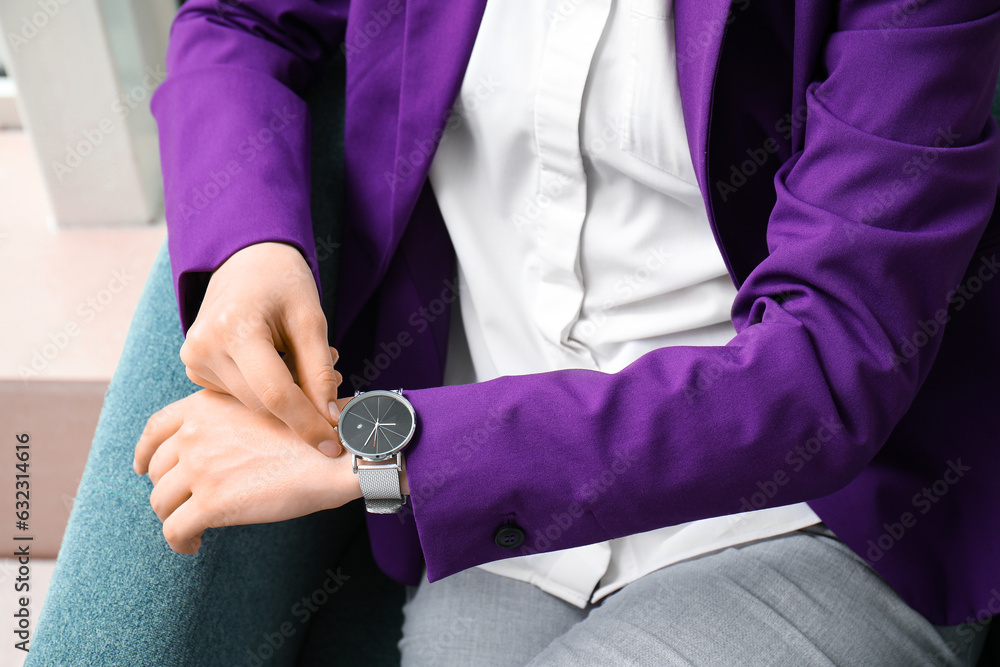 Young woman sitting on armchair and looking at stylish wristwatch in room, closeup