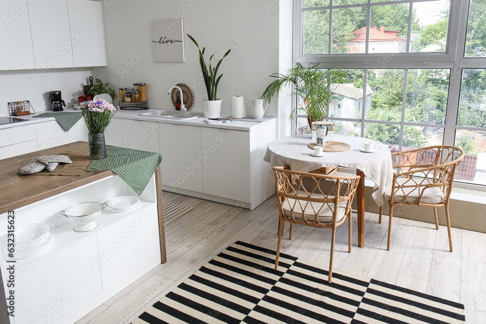 Interior of modern kitchen with white counters, island and dining table near big window