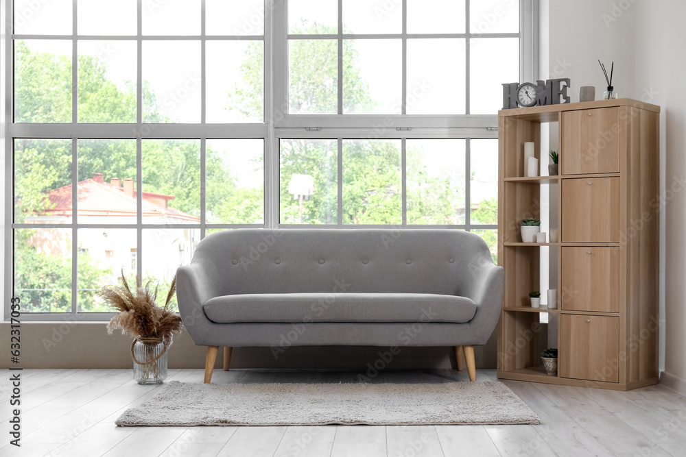 Interior of light living room with grey sofa and shelving unit near big window