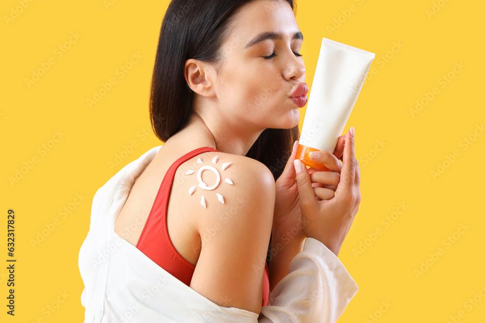 Young woman with sun made of sunscreen cream on her shoulder blowing kiss against yellow background,