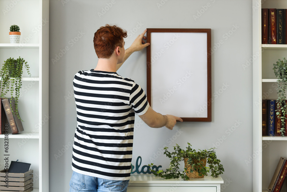 Young redhead man hanging blank frame on light wall at home, back view