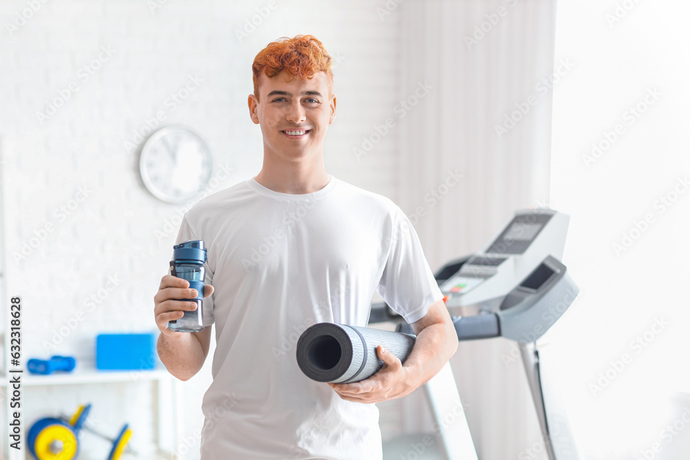 Young redhead man with sports water bottle and mat in gym