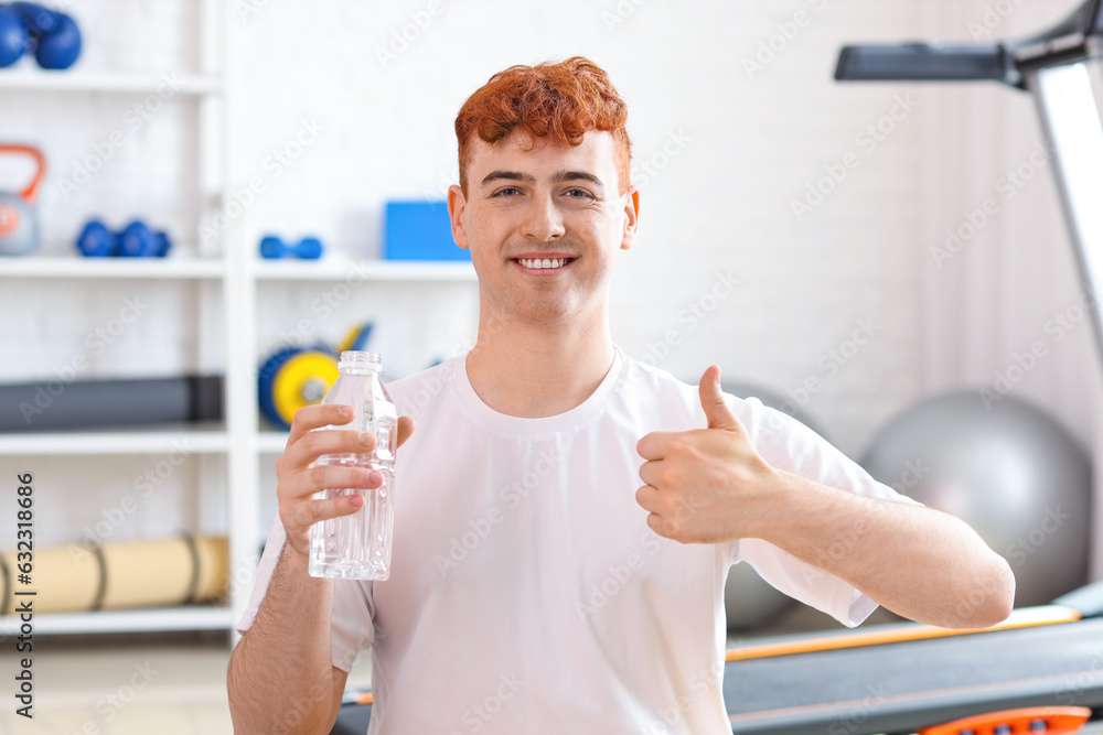 Young redhead man with bottle of water showing thumb-up in gym