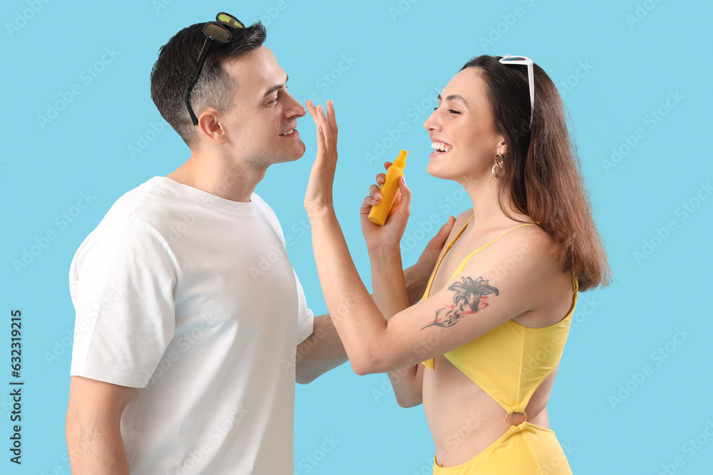 Young woman applying sunscreen cream onto her husbands face on blue background