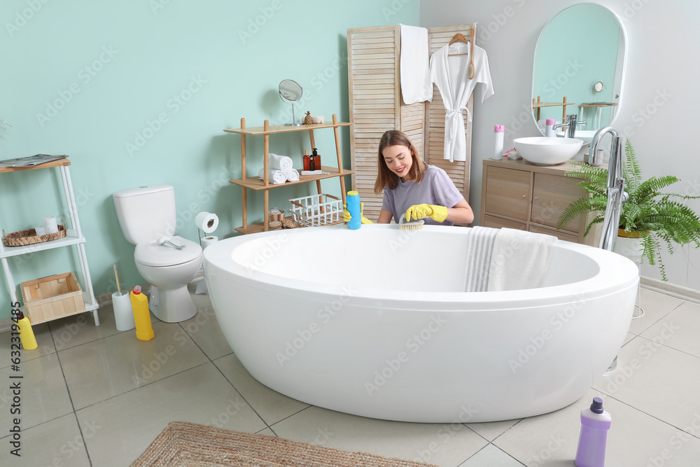 Young woman cleaning bathtub with brush in bathroom