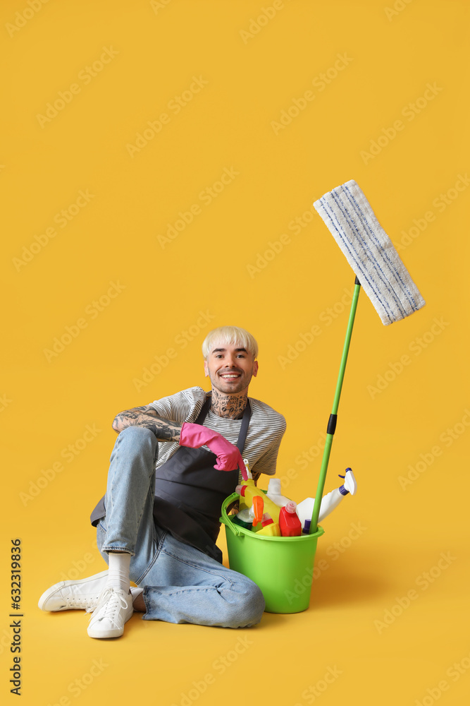 Young tattooed man with bucket of cleaning supplies sitting on orange background