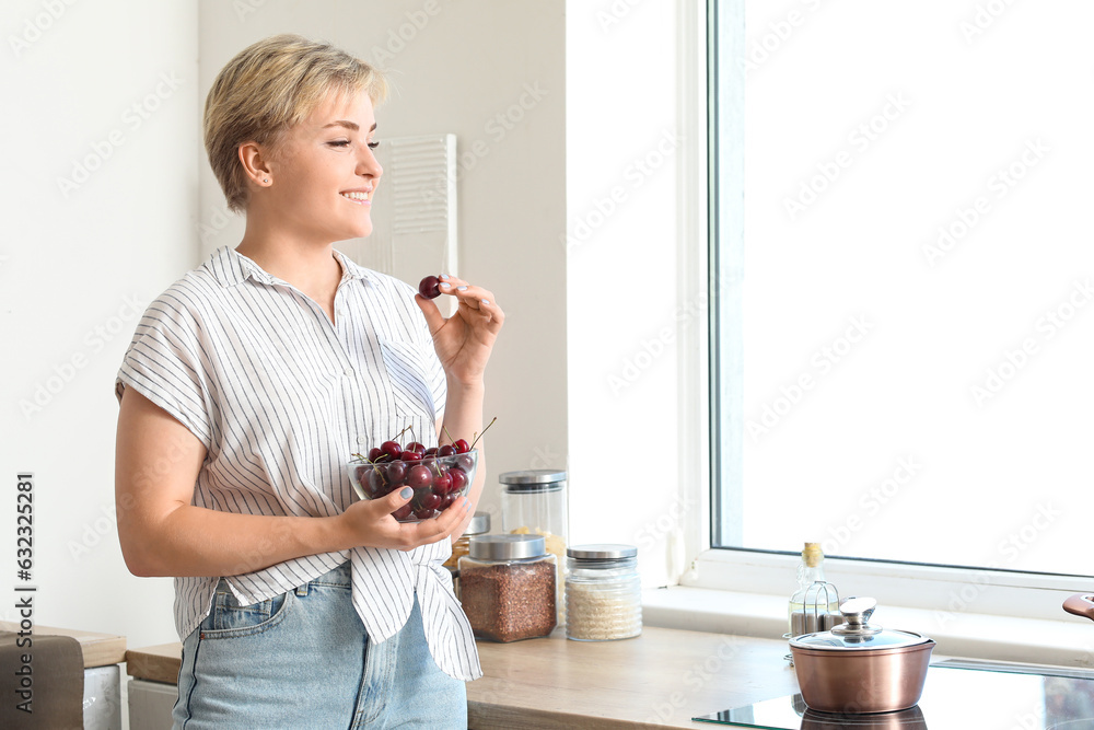 Beautiful young happy woman with bowl of ripe cherries in kitchen near window