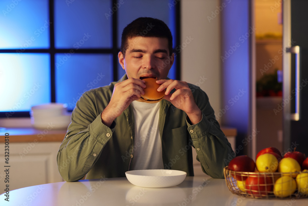 Hungry young man eating burger in kitchen at night