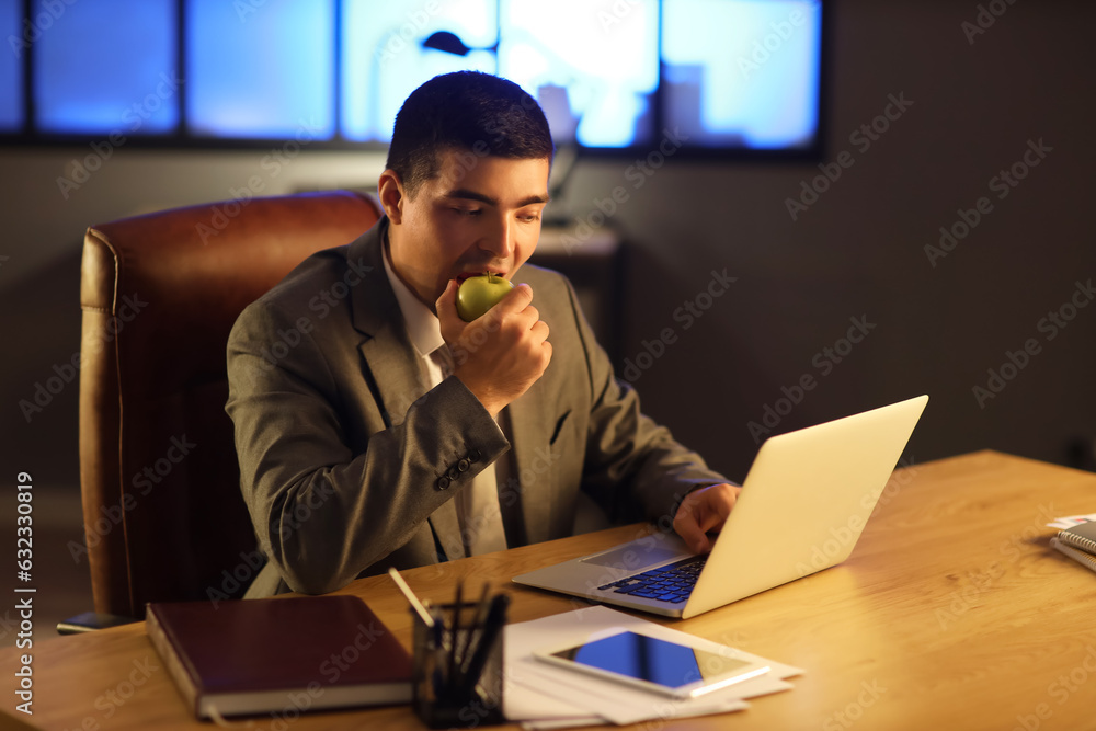 Young businessman eating apple in office at night