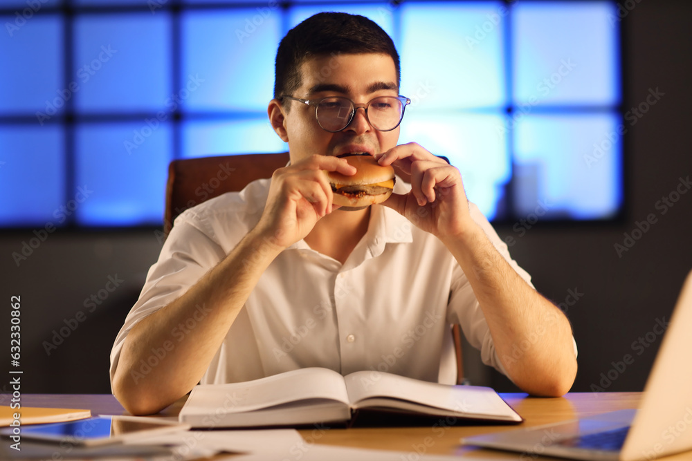 Young businessman eating tasty burger in office at night