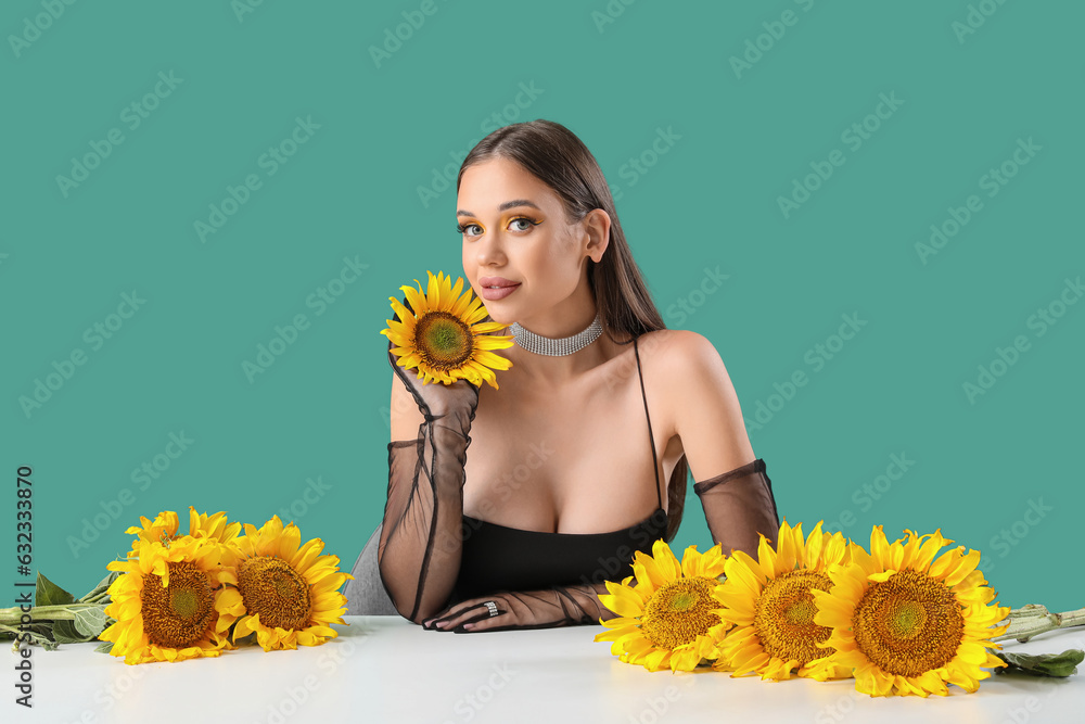 Elegant young woman with beautiful sunflowers sitting at table on green background