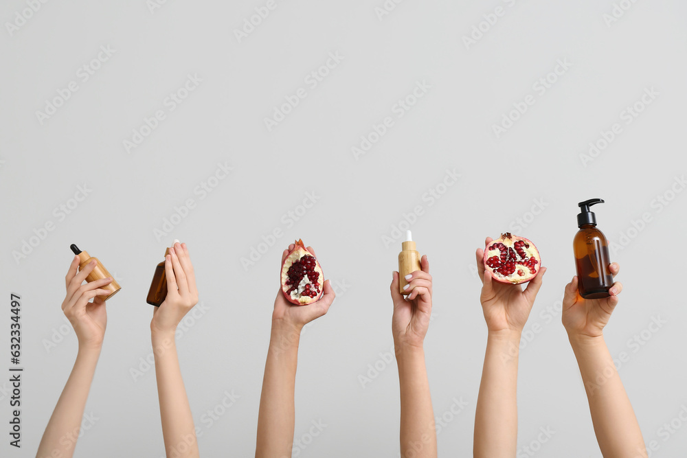 Female hands holding bottles with cosmetic products and pomegranate on grey background