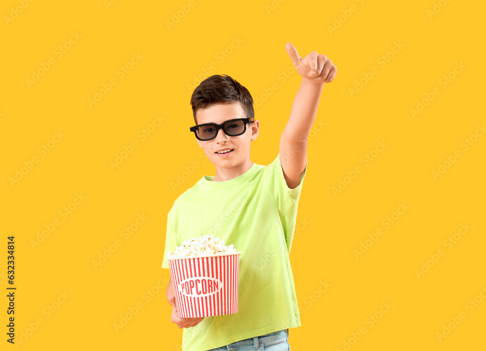 Cute little boy in 3D glasses with popcorn pointing at camera on orange background