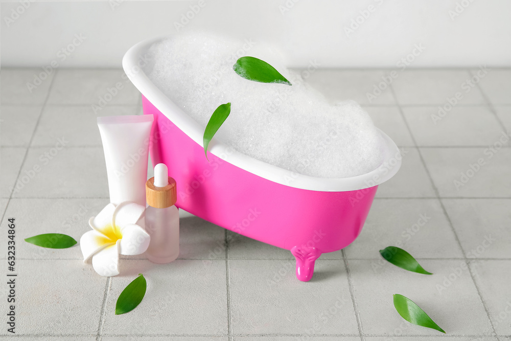 Small bathtub with foam, cosmetic products and plant leaves on light tile table