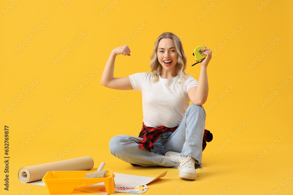 Young woman with measuring tape and wallpaper roll showing muscles on yellow background