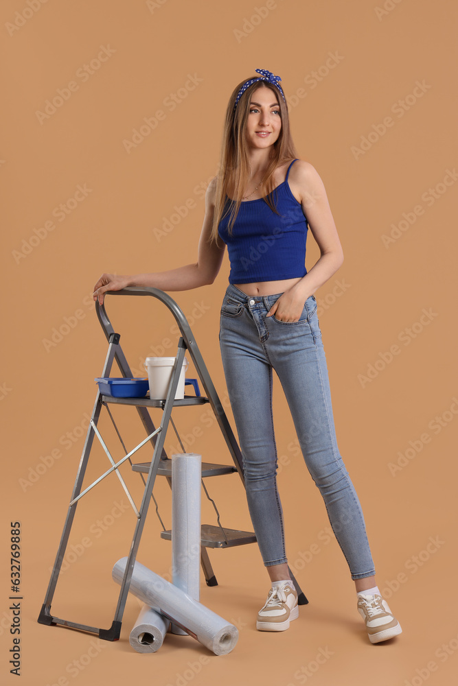 Young woman with wallpaper rolls, supplies and ladder on beige background