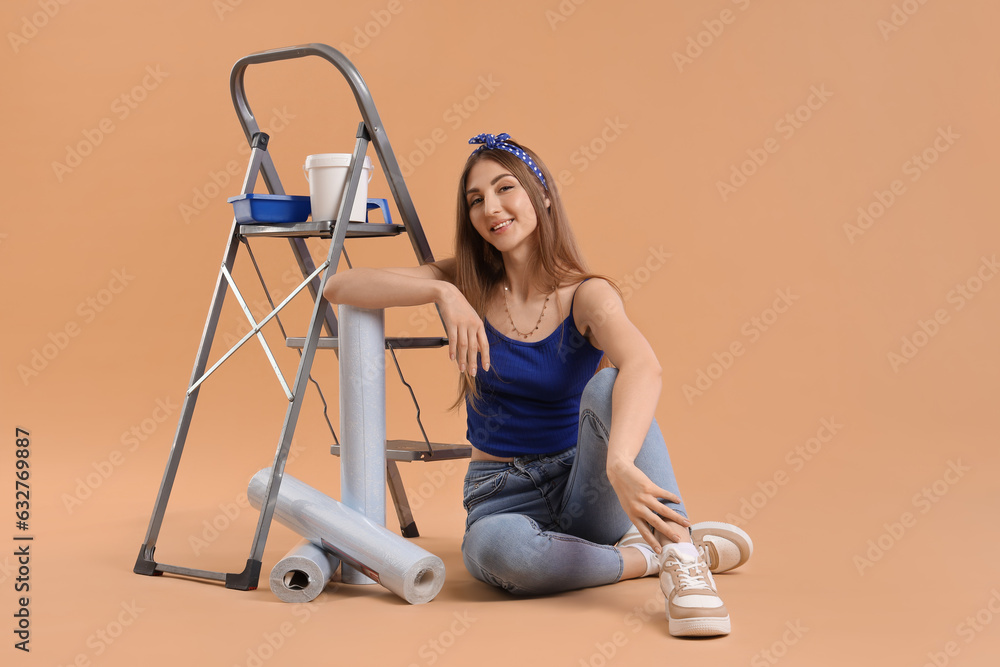 Young woman with wallpaper rolls, supplies and ladder on beige background