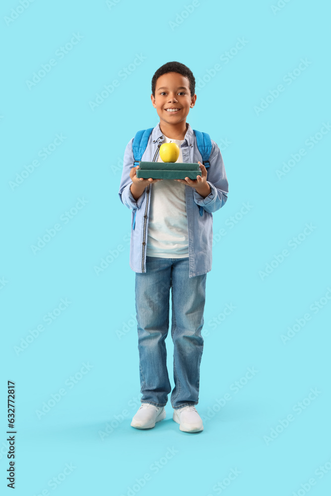 Little African-American schoolboy with books and apple on light blue background