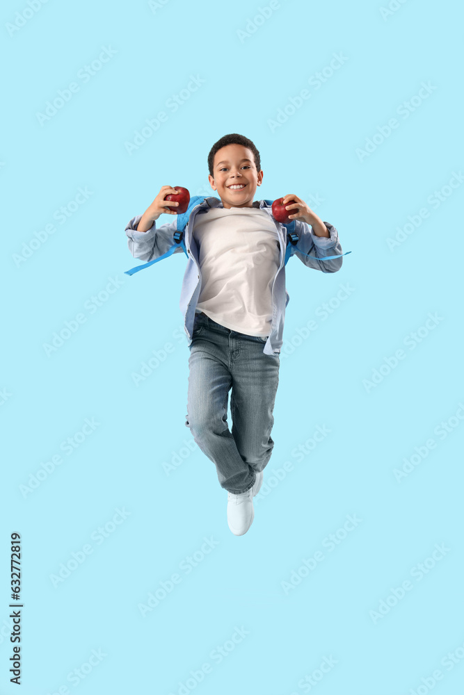 Jumping little African-American schoolboy with apples on light blue background