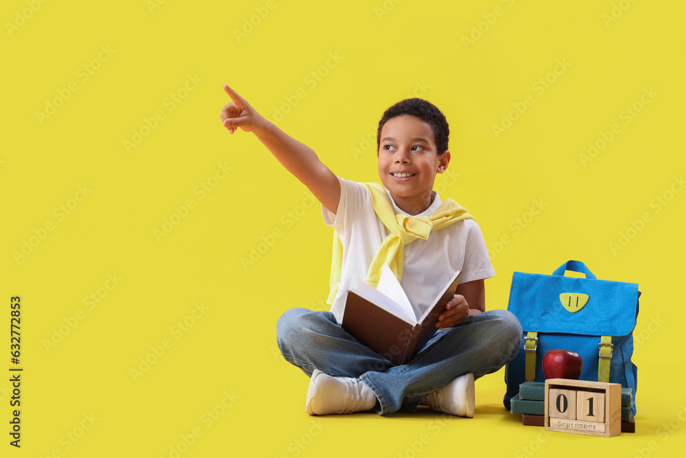 Little African-American schoolboy with book, backpack, calendar and apple on yellow background