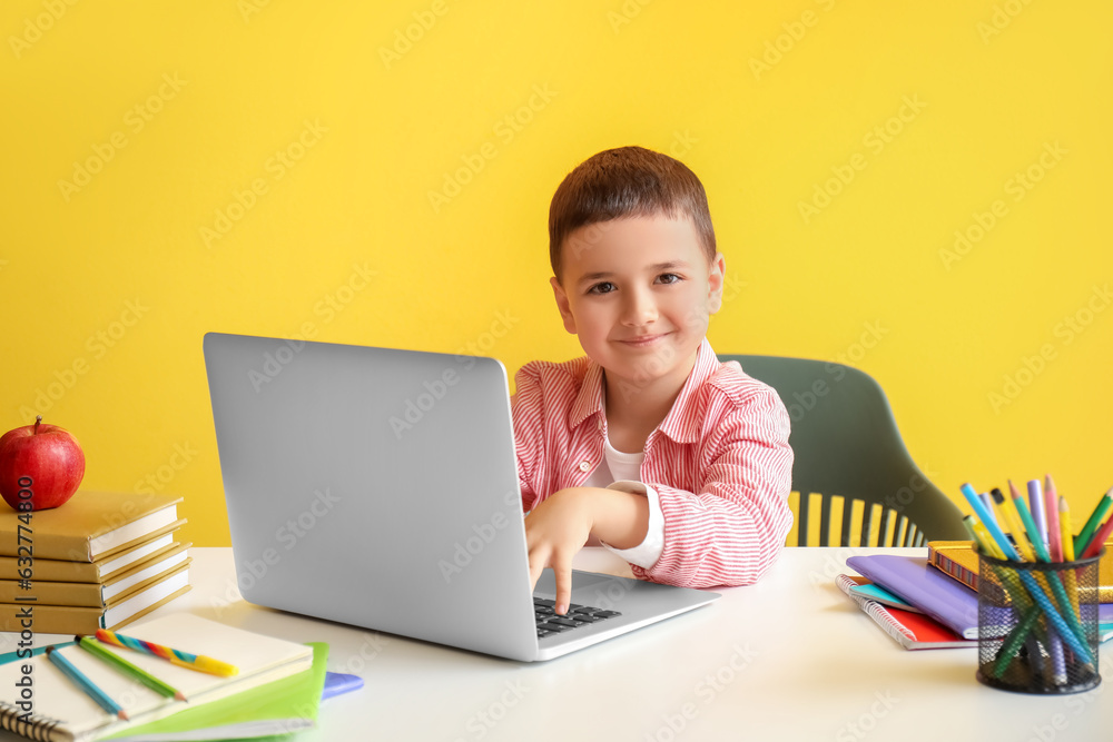 Cute little boy with laptop doing homework at table near yellow wall