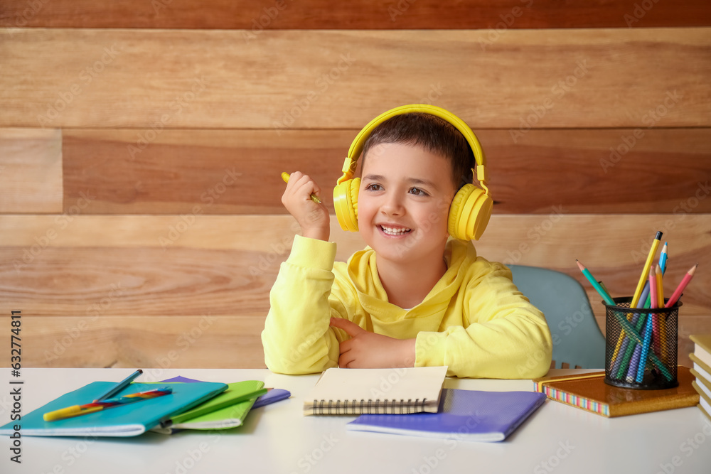 Cute little boy in headphones doing homework at table near wooden wall