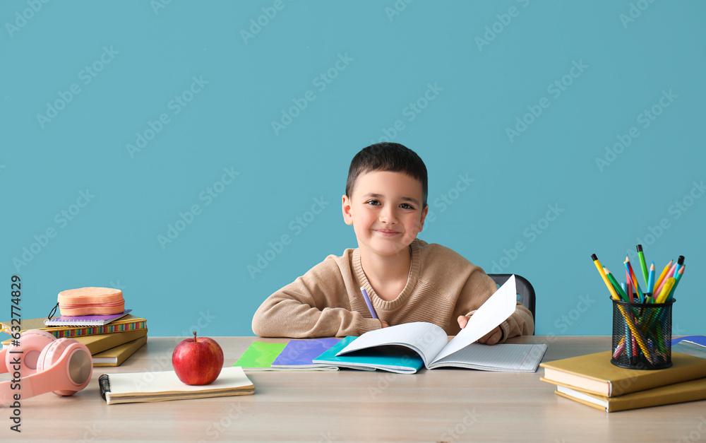 Cute little boy doing homework at table near blue wall