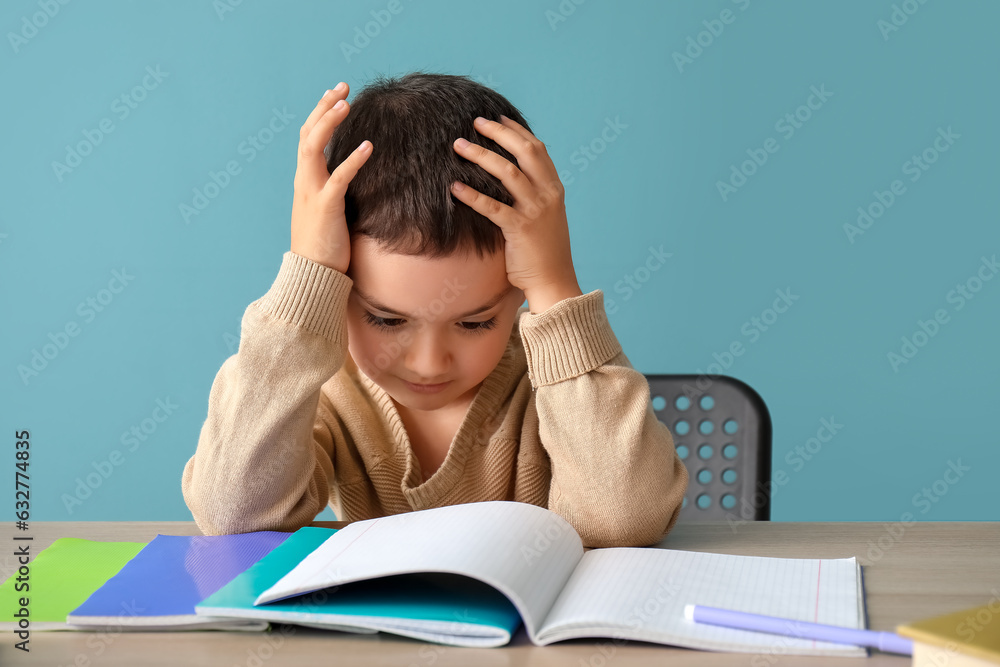 Stressed little boy doing homework at table near blue wall