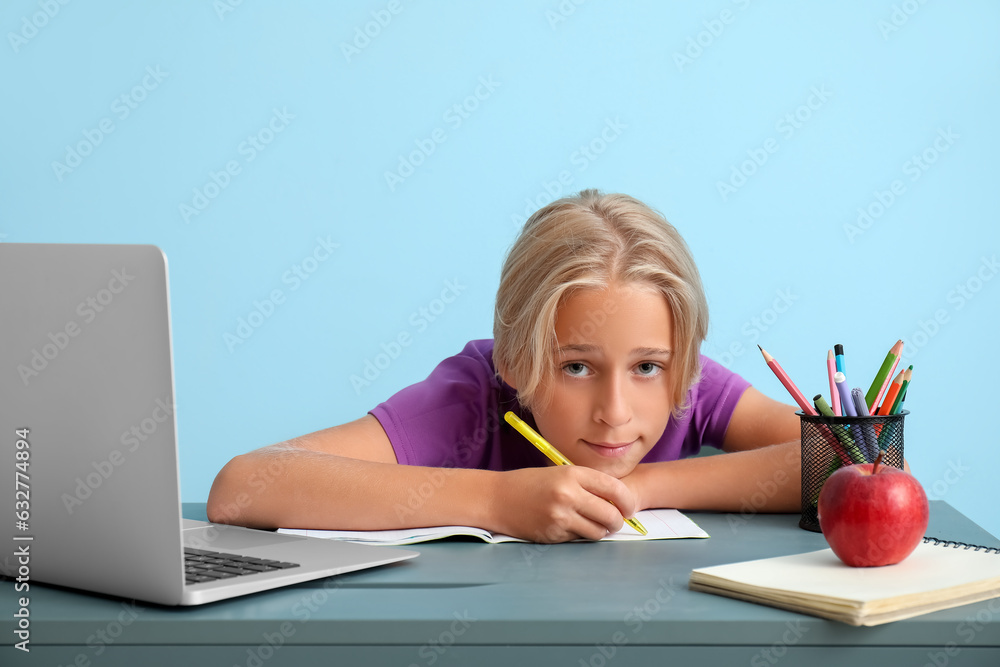 Tired little boy with laptop doing homework at table near blue wall