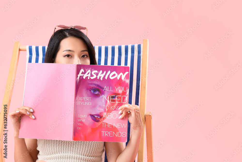 Beautiful Asian woman reading magazine in deck chair on pink background