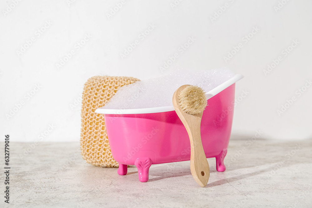 Small bathtub with soap foam, massage brush and sponge on table against light background