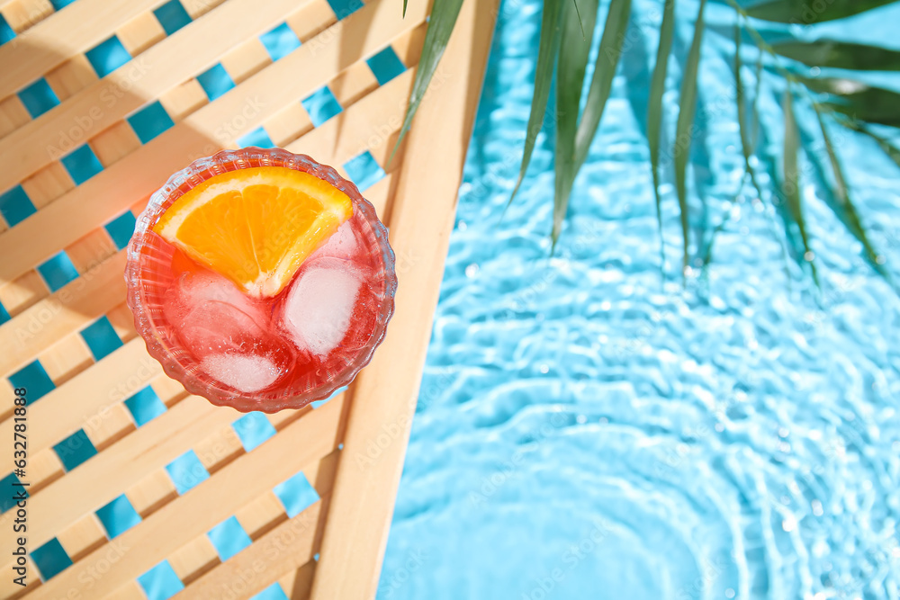 Glass of cold Negroni cocktail and palm leaf on wooden grid near swimming pool