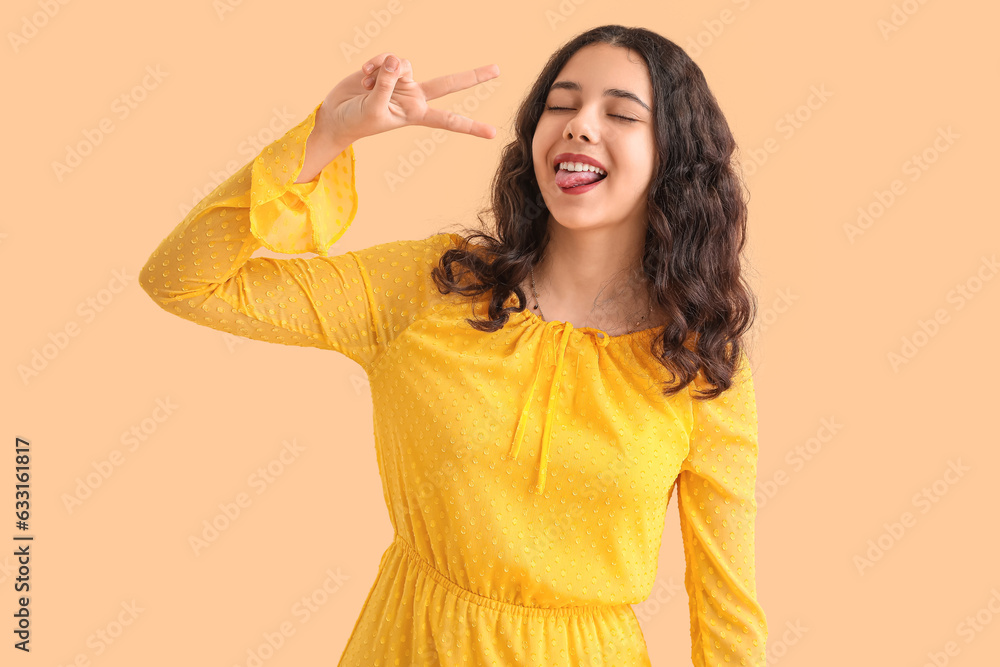 Teenage girl in yellow dress showing victory gesture on beige background