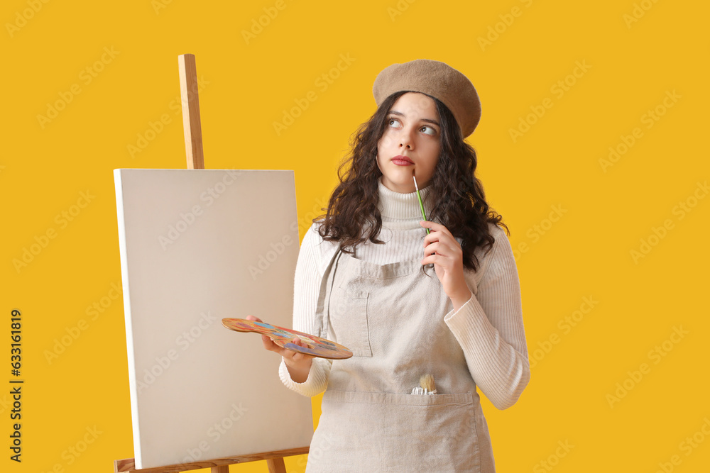 Thoughtful teenage artist with brush, paint palette and easel on yellow background