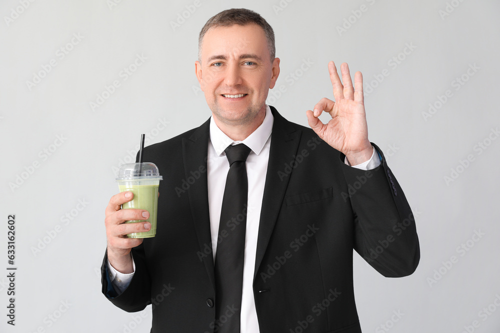 Mature businessman with glass of vegetable smoothie showing OK on light background