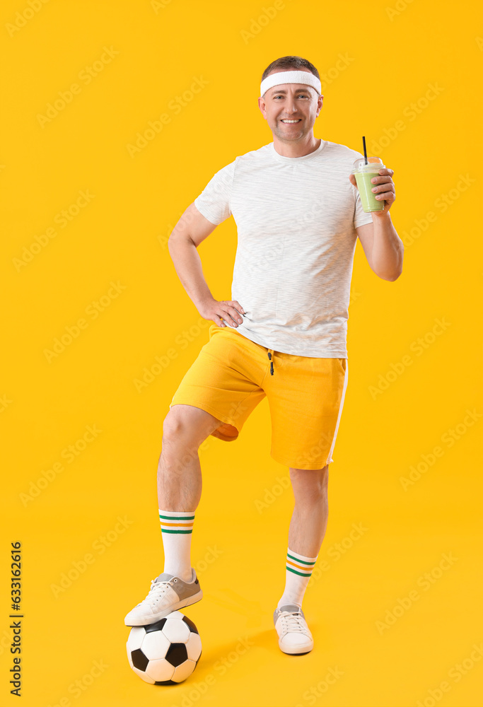 Sporty mature man with glass of vegetable smoothie and soccer ball on yellow background