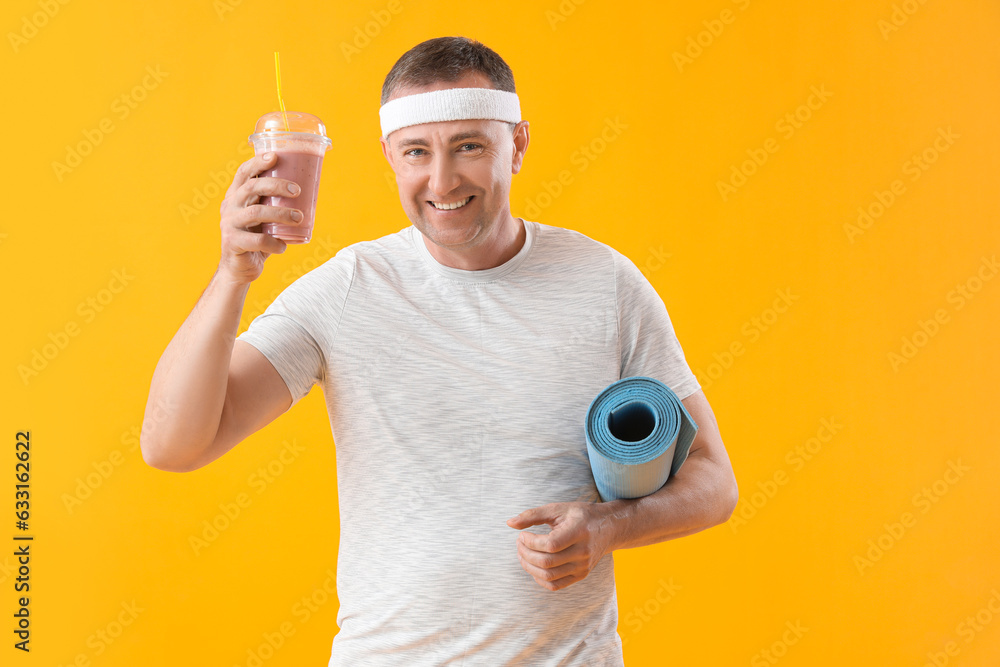 Sporty mature man with glass of fruit smoothie and fitness mat on yellow background