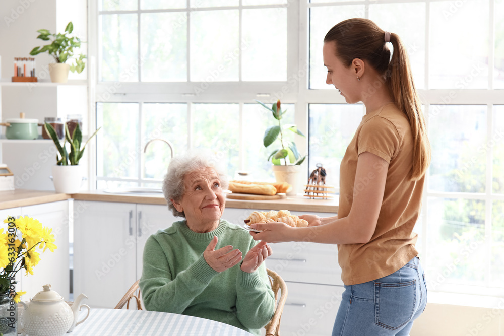 Young woman giving tasty croissants to her grandmother in kitchen