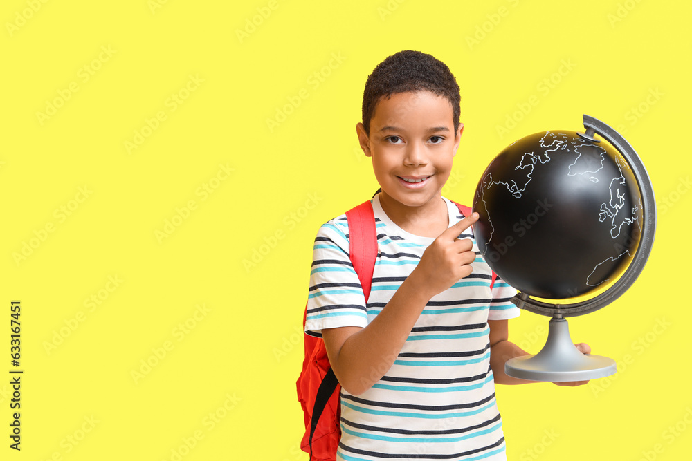 Little African-American boy with school backpack pointing at globe on yellow background