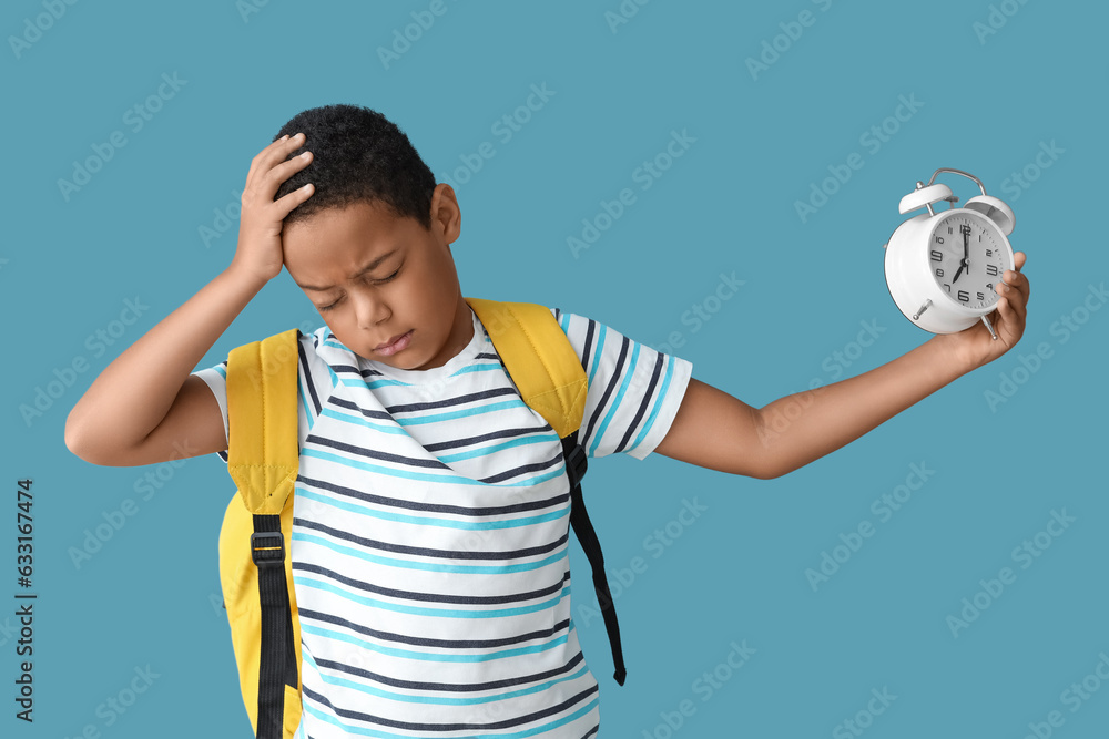 Tired African-American boy with school backpack and alarm clock on blue background