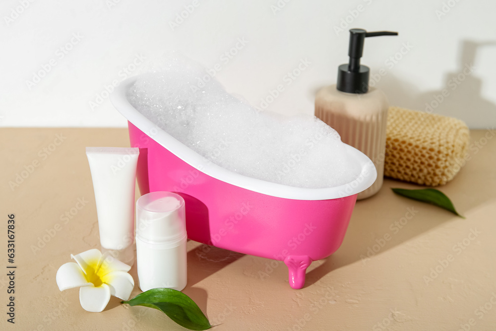 Small bathtub with foam, cosmetic products and sponge on color table against light background