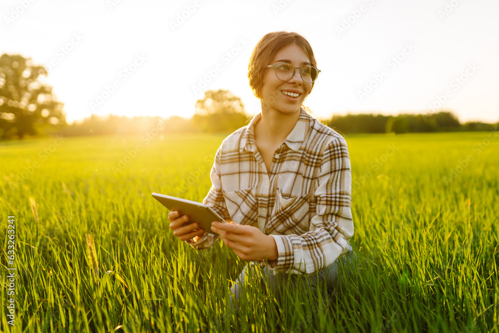 Young farmer woman with digital tablet evaluates shoots, green wheat sprouts in the field. Farm work