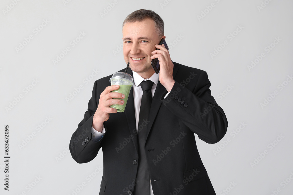 Mature businessman with glass of vegetable smoothie talking by mobile phone on light background