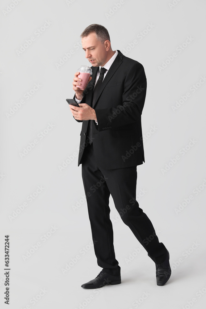 Mature businessman with mobile phone drinking fruit smoothie on light background