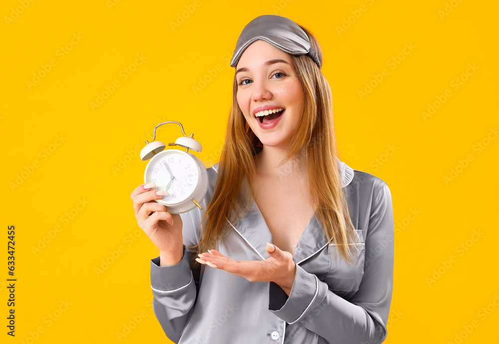 Young woman in pajamas with alarm clock on yellow background