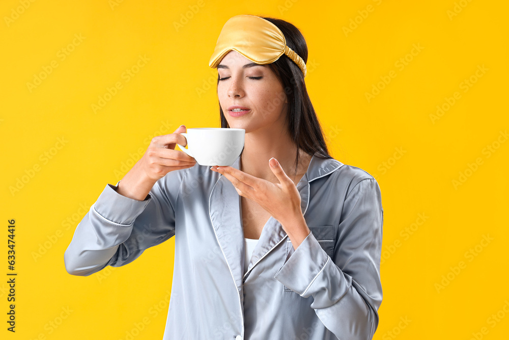 Young woman in pajamas with cup of coffee on yellow background