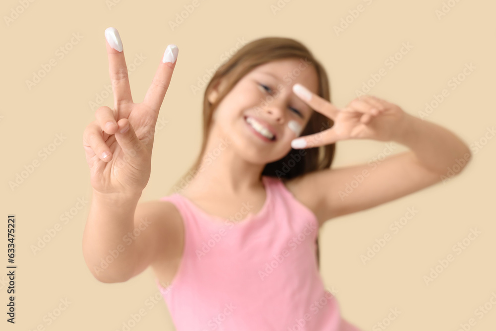 Little girl with sunscreen cream on her face showing victory gesture against beige background